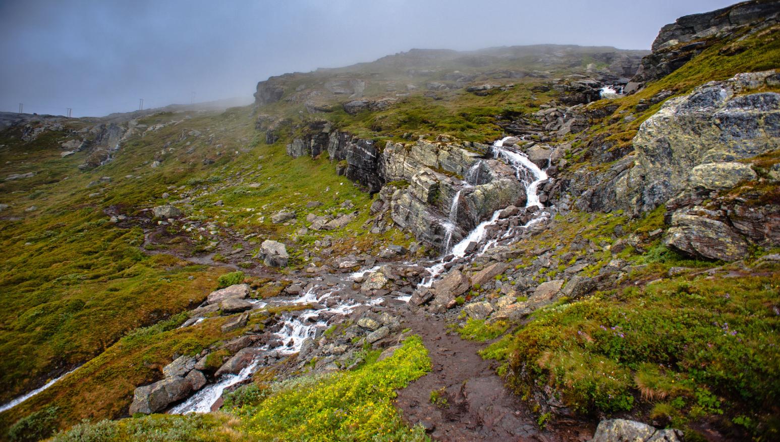 Small cascading stream flows through a rocky, green mountain landscape under a misty sky.