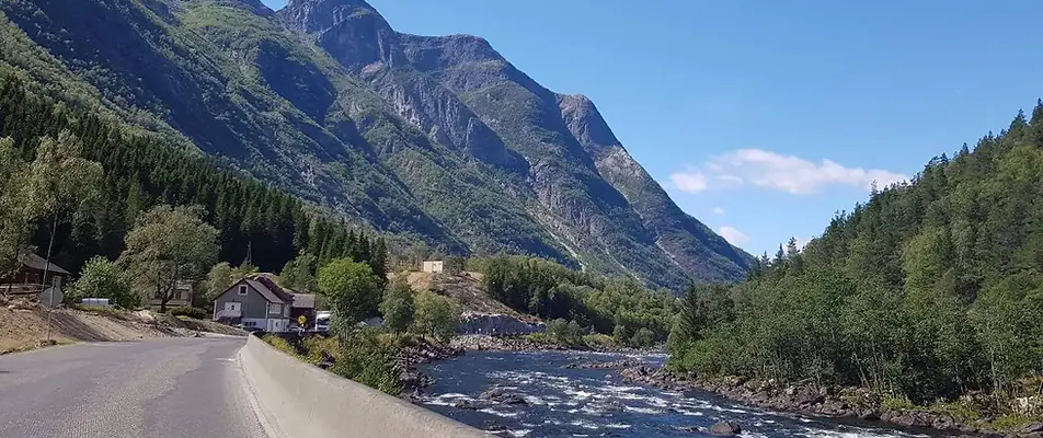 Scenic mountain landscape with a river flowing alongside a road, surrounded by lush green forests under a clear blue sky.