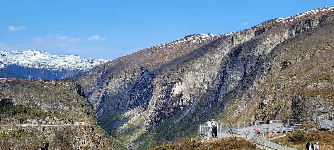Viewpoint with people overlooking a deep mountain valley, with snow-capped peaks in the distance under a clear sky.
