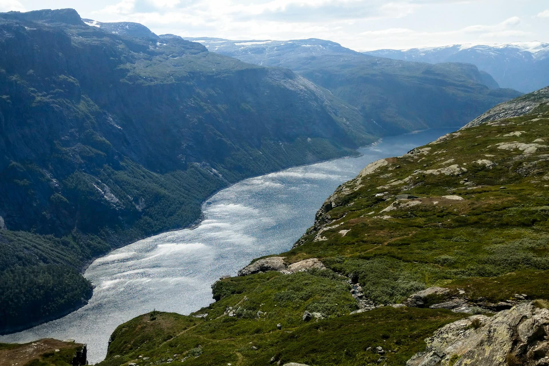 Panoramic view of a fjord winding between steep mountains, with rocky foreground and distant peaks.