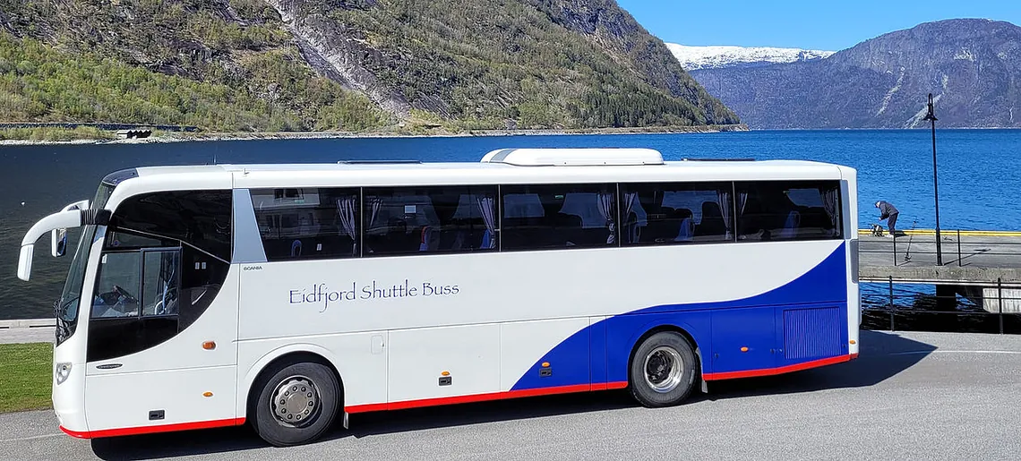 Hvit og blå turbuss merket "Eidfjord Shuttle Buss" parkert ved en fjord med fjell i bakgrunnen.