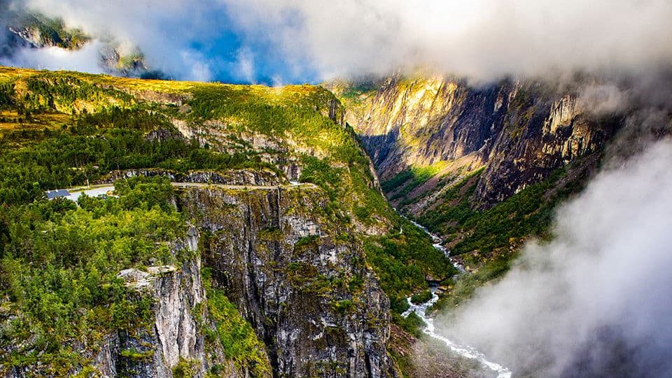 Majestic waterfall plunges into a deep canyon surrounded by cliffs, forest and low-hanging clouds.