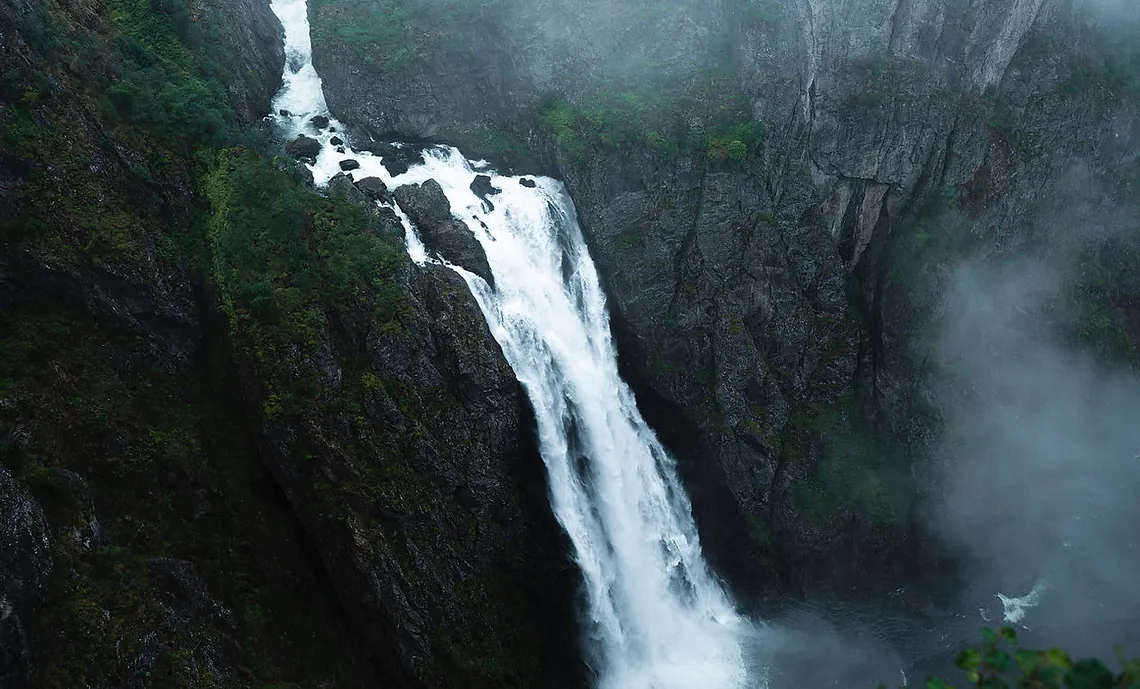 En foss stuper ned en bratt klippe, omgitt av tåke og grønn vegetasjon.