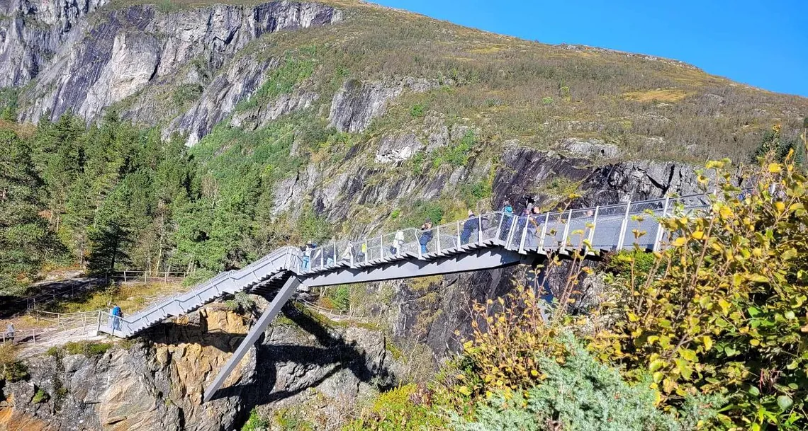 Gangbro over kløft med fargerik vegetasjon og blå himmel.